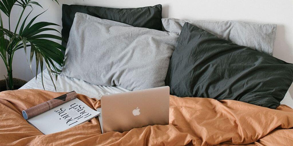 Laptop and magazine sitting atop bed with pillows and orange doonah